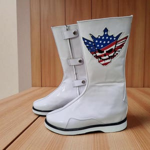 May include: Pair of white leather boots with a skull and American flag design. The boots have a mid-calf height, black soles, and a buckle detail. The design features a skull with a crown, wings, and the American flag.