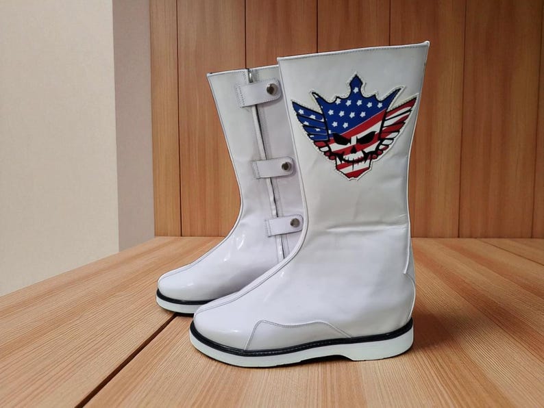 May include: Pair of white leather boots with a skull and American flag design. The boots have a mid-calf height, black soles, and a buckle detail. The design features a skull with a crown, wings, and the American flag.