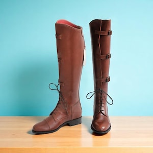 May include: Two brown leather riding boots. One boot has laces and the other has buckles. The boots are tall, reaching the knee, and have a reddish-brown hue. The background is a light blue.