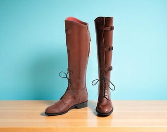 May include: Two brown leather riding boots. One boot has laces and the other has buckles. The boots are tall, reaching the knee, and have a reddish-brown hue. The background is a light blue.
