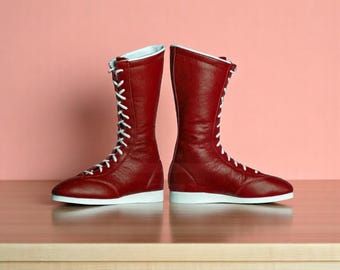 May include: Pair of tall, red leather boots with white laces and soles. The boots have a high-top design and a light blue interior trim. They are standing upright against a pink background.