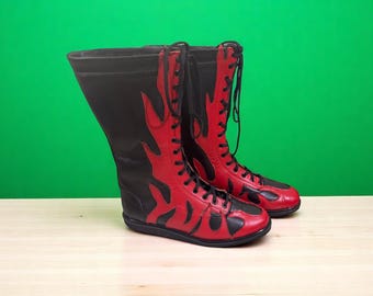 May include: A pair of red and black boots with a flame design. The boots are lace-up and have a high shaft. The boots are made of leather and have a black sole. The background is green.
