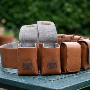 May include: A tan leather utility belt featuring multiple rectangular pouches with flap closures. Two pouches are open, revealing a grey interior lining, while others remain closed. Each pouch is secured with a small square of brown hook-and-loop fastener.