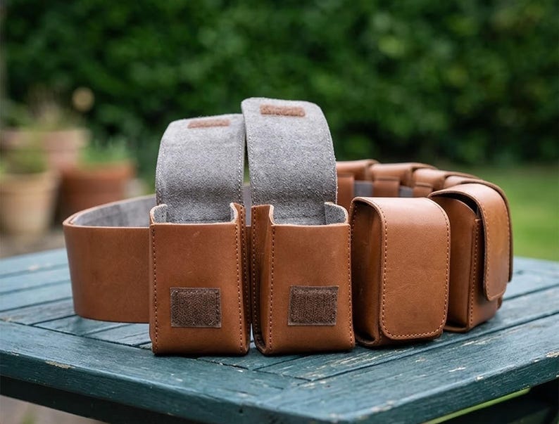 May include: A tan leather utility belt featuring multiple rectangular pouches with flap closures. Two pouches are open, revealing a grey interior lining, while others remain closed. Each pouch is secured with a small square of brown hook-and-loop fastener.
