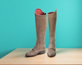 May include: A pair of tall, knee-high boots in a muted brown hue. The boots have a suede-like texture, with a contrasting pink interior visible at the top. Each boot features a strap with a buckle near the ankle. The boots are displayed on a light brown surface against a teal background.