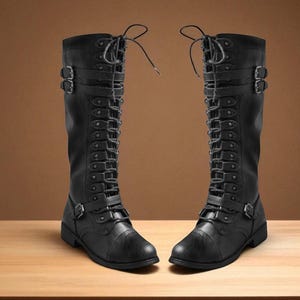 May include: Two black knee-high boots with multiple straps, buckles, and laces. The boots have a military or steampunk aesthetic, with a matte finish and a sturdy sole. The boots are displayed on a light brown wooden surface.