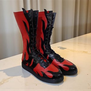 May include: A pair of knee-high boots with a striking flame design. The boots are primarily red with black accents, including the laces and the flame details. The boots have a flat sole and a glossy finish.