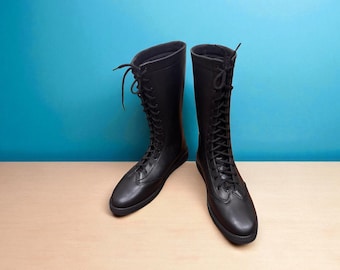 May include: A pair of black, knee-high boots with a lace-up design. The boots are made of a smooth material and have a flat sole. The boots are displayed against a light brown surface and a blue background.