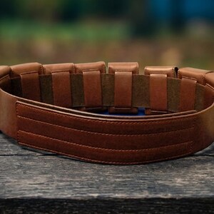 May include: A brown leather belt featuring a series of rectangular pouches attached along the exterior. The belt has a smooth, wide front panel with three horizontal stitched lines and a dark brown interior lining with multiple vertical loops.