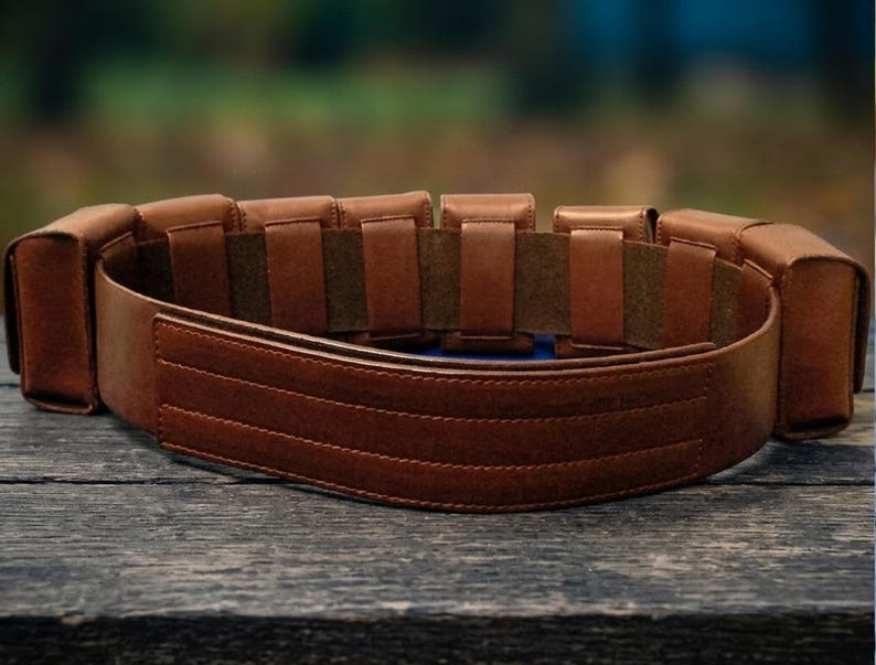 May include: A brown leather belt featuring a series of rectangular pouches attached along the exterior. The belt has a smooth, wide front panel with three horizontal stitched lines and a dark brown interior lining with multiple vertical loops.