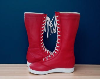 May include: A pair of bright red, knee-high boots with white laces. The boots have a white sole and a smooth, leather-like texture. The laces run up the front of the boots, creating a striking contrast against the red.