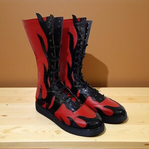May include: A pair of tall, red and black boots with a flame design. The boots have a lace-up front and a flat sole. The boots are made of a shiny material and are sitting on a wooden surface.