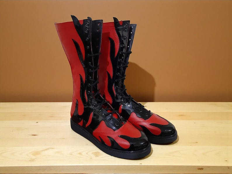 May include: A pair of tall, red and black boots with a flame design. The boots have a lace-up front and a flat sole. The boots are made of a shiny material and are sitting on a wooden surface.