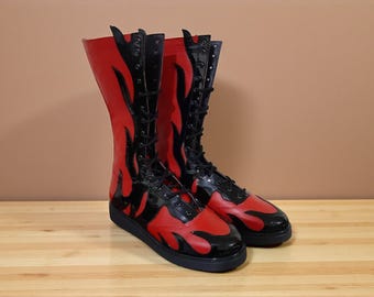 May include: A pair of red and black boots with a flame design. The boots are knee-high with black laces and a black sole. The boots are made of a shiny material and are displayed on a wooden surface.