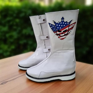 May include: Pair of white boots with a glossy finish, featuring a black sole and a decorative emblem. The emblem includes a skull, wings, and a crown, with red, white, and blue colors. The boots have buckles and are on a wooden surface.