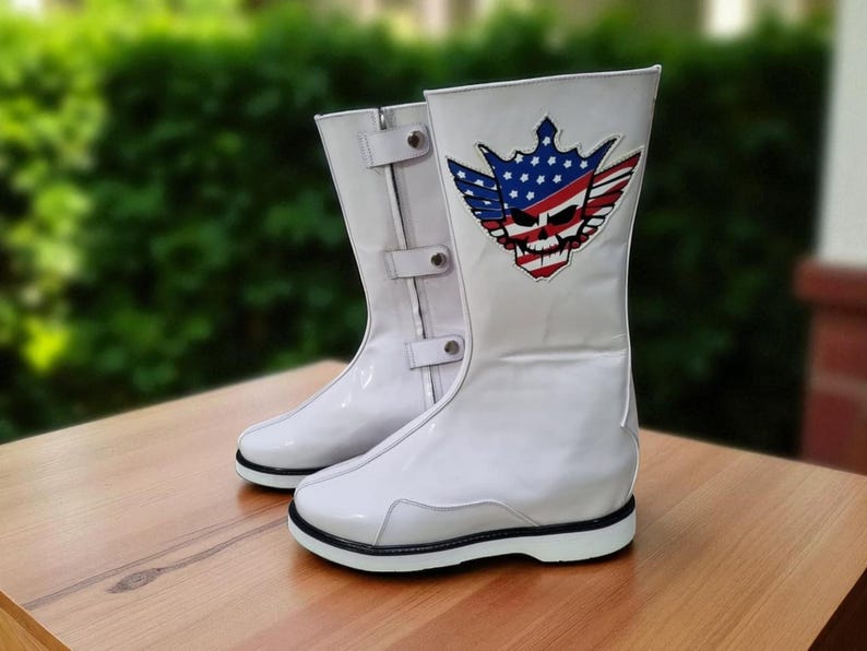 May include: Pair of white boots with a glossy finish, featuring a black sole and a decorative emblem. The emblem includes a skull, wings, and a crown, with red, white, and blue colors. The boots have buckles and are on a wooden surface.