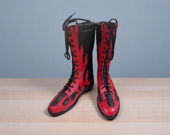 May include: A pair of red and black knee-high boots with a flame design. The boots have black laces and a black sole. The boots are made of leather and are on a light brown surface.