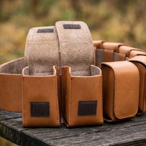 May include: A tan leather utility belt featuring multiple attached pouches with flap closures. Each pouch is secured with a dark brown hook-and-loop fastener. The belt is constructed from smooth, light brown leather with visible stitching along the edges.