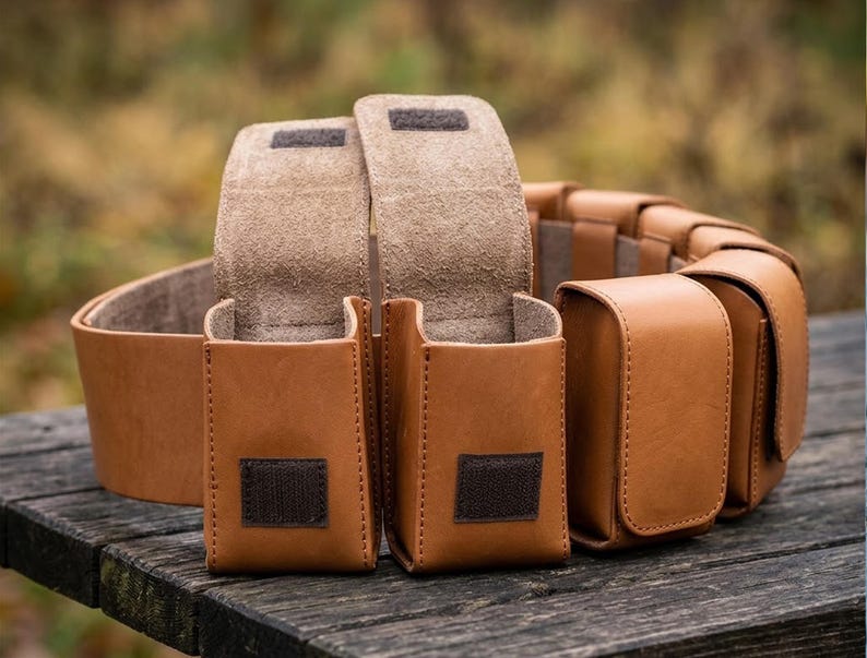 May include: A tan leather utility belt featuring multiple attached pouches with flap closures. Each pouch is secured with a dark brown hook-and-loop fastener. The belt is constructed from smooth, light brown leather with visible stitching along the edges.