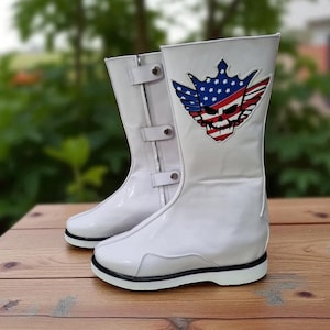 May include: Pair of white, high-top boots with a glossy finish. Each boot features a decorative patch with a skull, wings, and a crown design incorporating the American flag. The boots have black soles and adjustable straps.