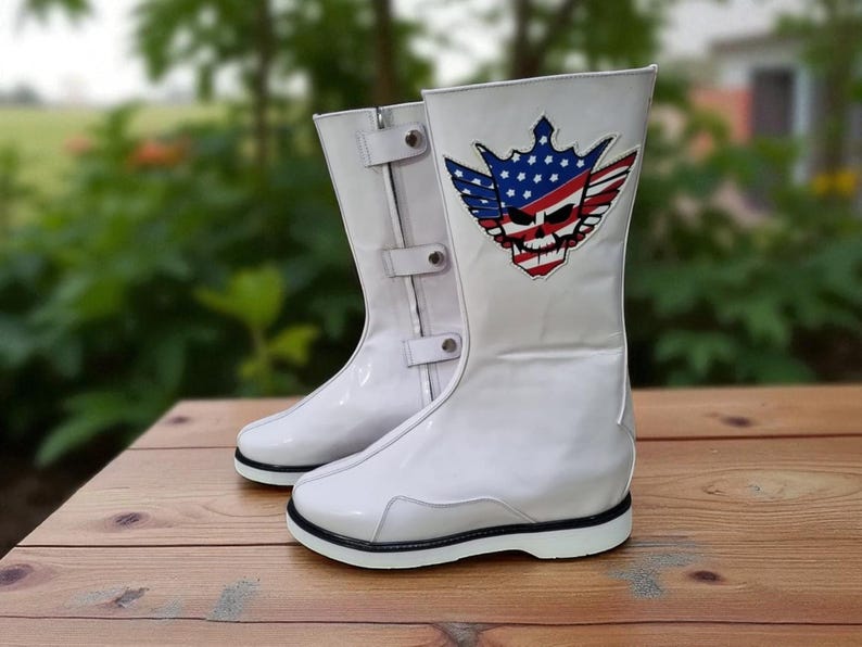 May include: Pair of white, high-top boots with a glossy finish. Each boot features a decorative patch with a skull, wings, and a crown design incorporating the American flag. The boots have black soles and adjustable straps.