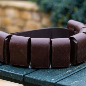 May include: A dark brown leather utility belt featuring multiple rectangular pouches attached along the length of the strap. Each pouch has a flap closure and a smooth, consistent texture, creating a structured and functional accessory design.
