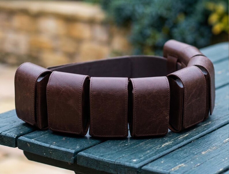 May include: A dark brown leather utility belt featuring multiple rectangular pouches attached along the length of the strap. Each pouch has a flap closure and a smooth, consistent texture, creating a structured and functional accessory design.