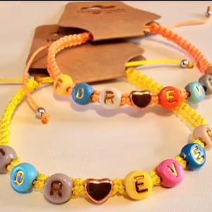 Può includere: Due braccialetti dell'amicizia regolabili. Uno è giallo con la parola "FOREVER" scritta con perline colorate. L'altro è arancione e giallo con la parola "LOVE". Ogni braccialetto ha un ciondolo a forma di cuore.