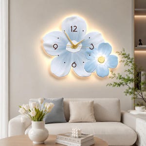 Puede incluir: Un reloj de pared con forma de dos flores superpuestas de color azul claro con bordes blancos. La flor más grande tiene manecillas doradas y números marrones oscuros para el 3, 6, 9 y 12. La flor pequeña tiene un centro amarillo. Luz cálida.