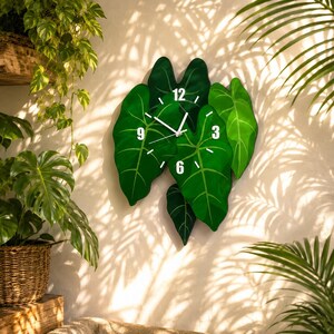 May include: A wall clock shaped like a cluster of overlapping green alocasia leaves. The clock face features white hour and minute hands, with the numbers 3, 6, 9, and 12 printed in white on the leaves. The design mimics natural foliage.