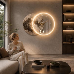 May include: A circular wall clock featuring a transparent face with gold-coloured hands and a glowing warm LED ring. The clock is mounted over a textured, moon-like circular wall art piece with dark and light grey mottled patterns.