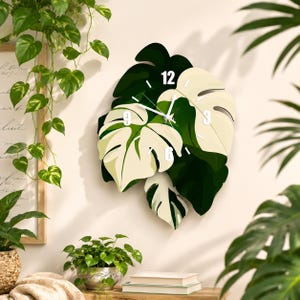 Peut inclure: Une horloge murale en forme de groupe de feuilles de monstera dans des tons vert foncé et crème. Le cadran présente des chiffres blancs aux positions 12, 3, 6 et 9, avec de fines aiguilles blanches pour les heures, minutes et secondes.