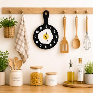 May include: A round black wall clock shaped like a cutting board with a central fried egg graphic, white numbers from 1 to 12, and black hands. The clock hangs on a wooden rack alongside a checkered towel, a wooden spatula, a spoon, and a whisk.