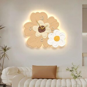 Puede incluir: Un reloj de pared con forma de dos flores superpuestas en tonos beige claro y blanco. La flor más grande tiene un centro marrón oscuro y manecillas doradas, mientras que la flor más pequeña tiene un centro amarillo. El reloj emite luz.
