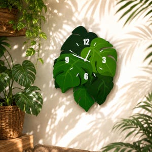 Pode incluir: Um relógio de parede em forma de um grupo de folhas de monstera sobrepostas em vários tons de verde. O mostrador do relógio apresenta ponteiros brancos para as horas e minutos, com os números 3, 6, 9 e 12 impressos em branco sobre as folhas.