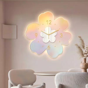Puede incluir: Un reloj de pared con forma de flor que presenta un degradado pastel de rosa, amarillo y azul. El reloj tiene un centro blanco, manecillas doradas y números dorados en las posiciones 12, 3, 6 y 9. Los bordes emiten un brillo suave y cálido.