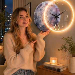 May include: A circular wall clock featuring a dual-layered design with a textured, abstract blue and purple painted disc partially overlapped by a clear glass face illuminated by a warm LED ring, displaying black hands and gold numerals 12, 6, and 9.