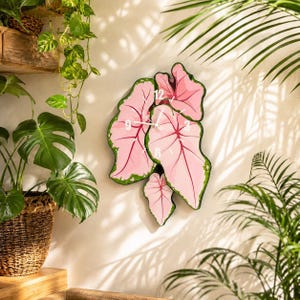 Peut inclure: Une horloge murale en forme de groupe de feuilles de Caladium roses avec des bords verts et des nervures rose foncé. Le cadran présente des chiffres blancs aux positions 12, 3, 6 et 9, avec de fines aiguilles blanches centrées sur les feuilles.