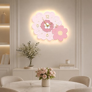 May include: A wall clock shaped like two overlapping flowers in shades of light and dark pink. The clock face features gold-colored hands and numerals at the 12, 3, 6, and 9 positions. A soft, warm light glows from behind the floral structure.