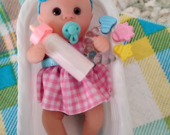 May include: A small baby doll with a light skin tone, wearing a pink and white gingham dress and a blue yarn headband. The doll has a light blue pacifier, holds a pink baby bottle, and has a multi-colored plastic rattle with star and shape charms.