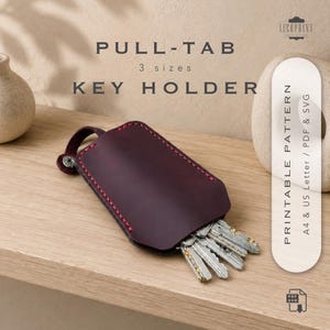 Pode incluir: Um porta-chaves de couro roxo escuro com costuras vermelhas e um botão de pressão de metal prateado. O porta-chaves contém um conjunto de chaves de metal. O texto na imagem diz: PULL-TAB 3 sizes KEY HOLDER PRINTABLE PATTERN A4 & US Letter / PDF & SVG.