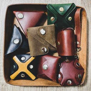 May include: Assortment of nine leather coin purses in various colors and designs, including green, brown, black, and yellow. The purses are arranged in a tray.