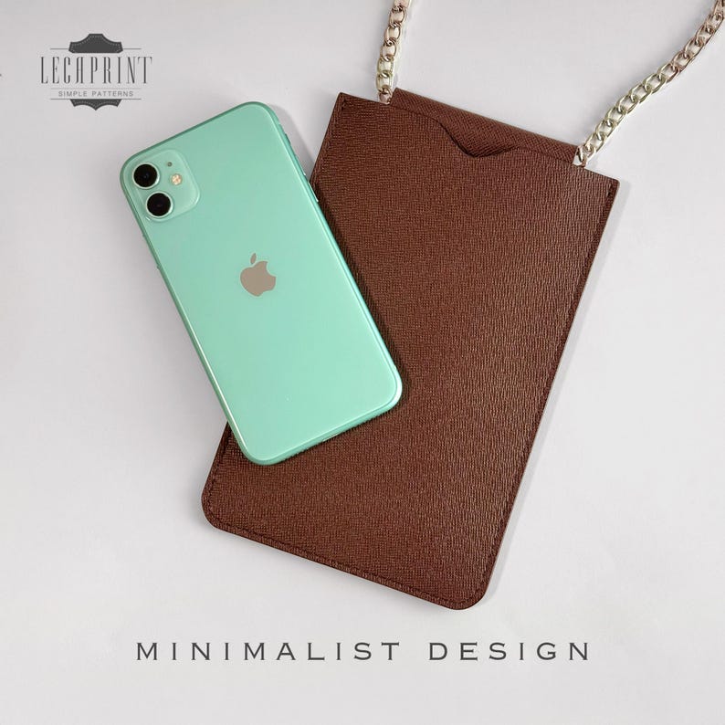 K&ouml;nnte beinhalten: Eine braune Leder-Handyh&uuml;lle mit einem silbernen Kettenriemen. Die H&uuml;lle hat eine Tasche auf der R&uuml;ckseite f&uuml;r ein Telefon und der Text "MINIMALIST DESIGN" ist unter der H&uuml;lle aufgedruckt. Ein mintgr&uuml;nes iPhone ist in der H&uuml;lle zu sehen.