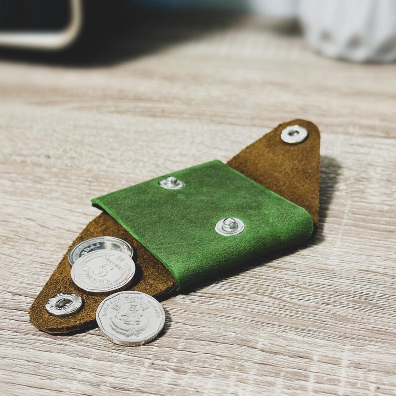 Pu&ograve; includere: Un porta monete in pelle verde con una patta in pelle marrone. Il porta monete &egrave; aperto e contiene diverse monete d'argento.