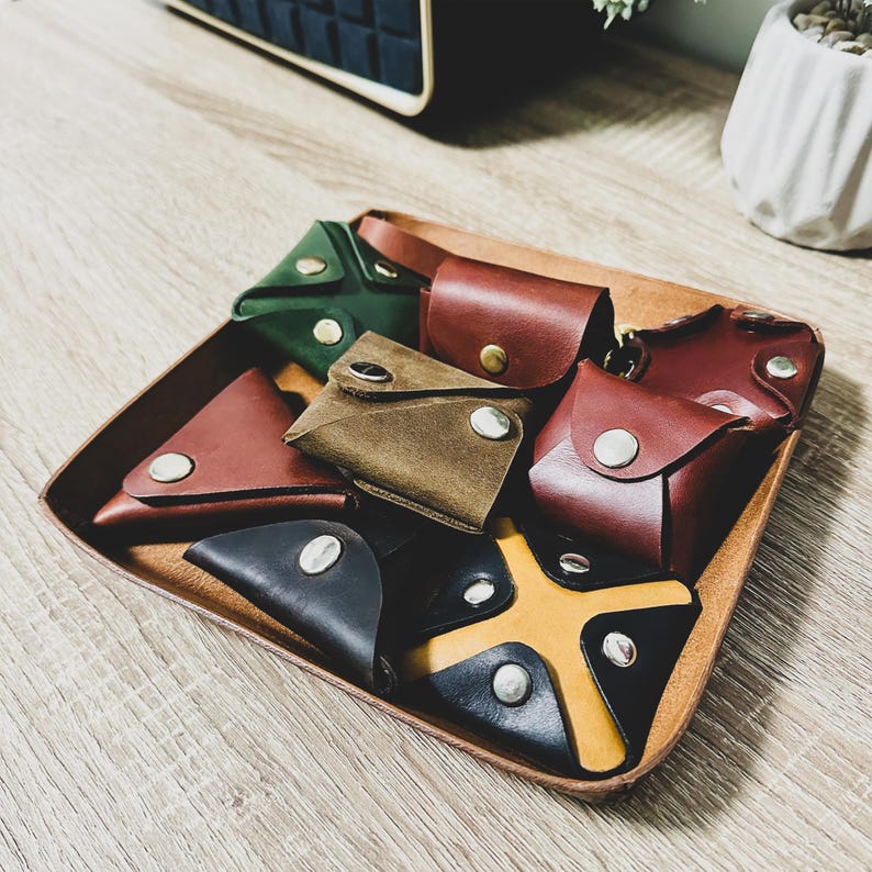 May include: A collection of eight small leather wallets in various colors, including green, brown, red, and black, arranged on a wooden surface. The wallets are made of different types of leather and have different designs.