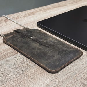 May include: A distressed brown leather phone case with a silver chain strap. The case has a flap closure with a snap and is next to a black laptop on a wooden surface. The leather has a worn, textured appearance.