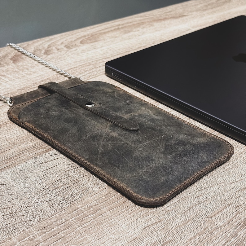 May include: A distressed brown leather phone case with a silver chain strap. The case has a flap closure with a snap and is next to a black laptop on a wooden surface. The leather has a worn, textured appearance.