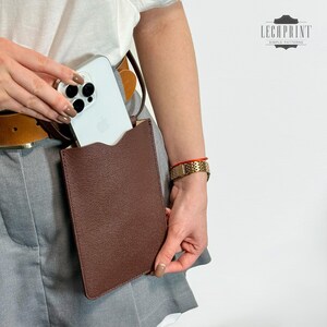 K&ouml;nnte beinhalten: Ein braunes Leder-Handy-Etui mit einem wei&szlig;en iPhone darin. Das Etui ist an einem braunen G&uuml;rtel mit einem Riemen befestigt. Das Etui wird von einer Person getragen, die ein wei&szlig;es Hemd und eine graue Hose tr&auml;gt.
