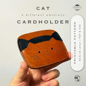 Puede incluir: Un tarjetero de cuero marrón con un diseño de cara de gato. Las orejas del gato son negras y la cara tiene detalles cosidos para los ojos y los bigotes. El texto "CAT" y "CARDHOLDER" están en la parte superior. El tarjetero se sostiene en una mano.