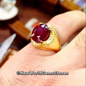 May include: A gold ring with a large, faceted, deep red gemstone, possibly a ruby, set in a decorative bezel with small, clear accent stones. The ring has a thick band and is displayed on a hand. The text "@NewWorldGemsStones" is visible.